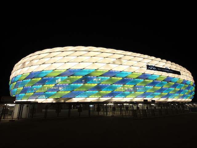 Allianz Arena in Munich illuminated in blue, green and white at night, with UEFA Champions League branding on its exterior.