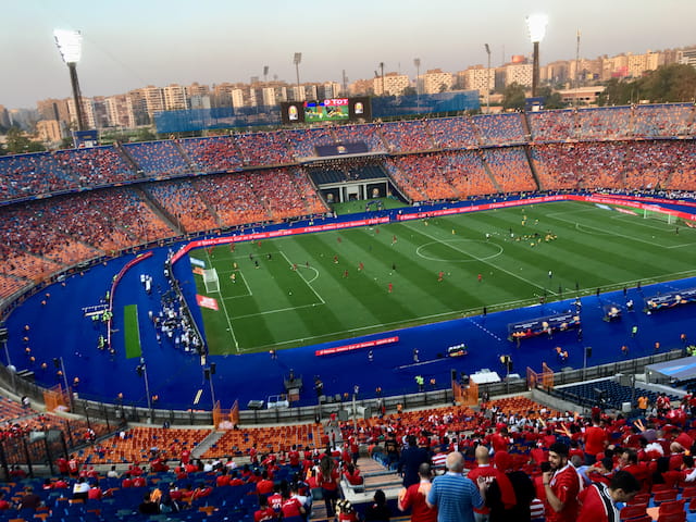 Wide view of Cairo International Stadium, showing the football pitch, running track and red stands.