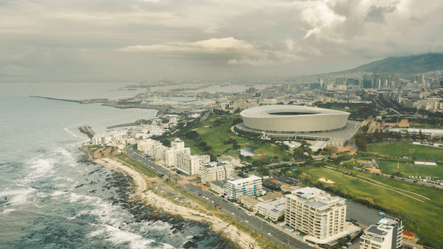 Aerial view of DHL Stadium beside Cape Town’s coastline, with the city and mountains beneath dramatic clouds.