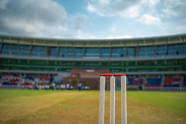 Three white cricket stumps topped with red bails, with a stadium blurred in the background.