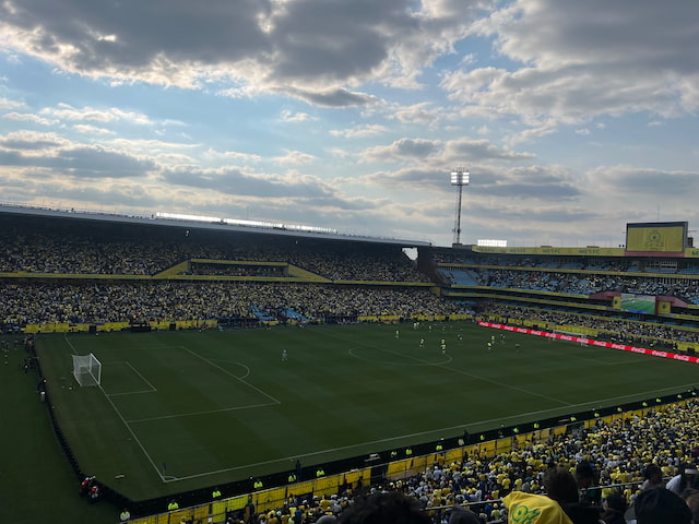 Mamelodi Sundowns supporters in yellow at Loftus Versfeld, with players on the pitch beneath a cloudy sky.